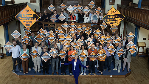 Daisy Cooper MP is standing in front of a crowd of Lib Dem activists, smiling and looking at the camera. The activists are holding orange diamond shaped Liberal Democrat signs.