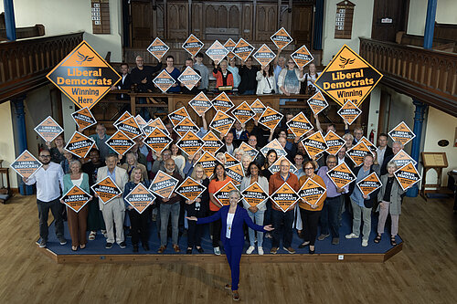 Daisy Cooper MP is standing in front of a crowd of Lib Dem activists, smiling and looking at the camera. The activists are holding orange diamond shaped Liberal Democrat signs.