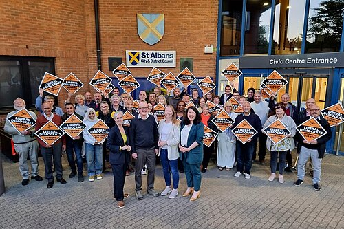A group of people are standing in front of the St Albans district council offices. Four people are stood in a line at the front (Daisy, Paul, Helen and Victoria). All of them are smiling and looking at the camera. There is a crowd stood behind them, all holding orange diamonds displaying the words Liberal Democrats and the Liberal Democrat bird logo.
