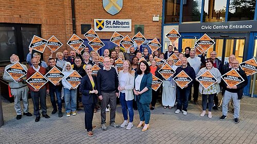 A group of people are standing in front of the St Albans district council offices. Four people are stood in a line at the front (Daisy, Paul, Helen and Victoria). All of them are smiling and looking at the camera. There is a crowd stood behind them, all holding orange diamonds displaying the words Liberal Democrats and the Liberal Democrat bird logo.