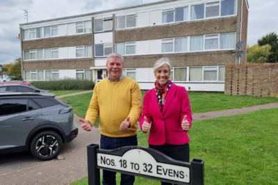 Two people (Tony on the left, Daisy on the right) are standing in front of a block of flats, and behind a sign displaying flat numbers. They are both smiling, looking directly at the camera, and giving a thumbs up.