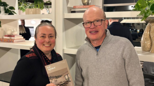 Paul and Jacqui are both smiling and standing in front of a bookcase, at the St Albans Co-Space opening event. Jacqui is holding a Co-Space leaflet.