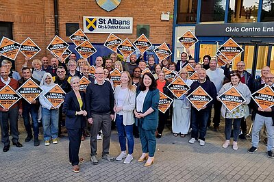 A group of Lib Dem councillors are standing in front of the St Albans District Council building, holding diamond shaped Liberal Democrat placards. Daisy Cooper MP, Councillor Paul de Kort, Councillor Helen Campbell and Victoria Collins MP (from left to right) are standing in front of the group.