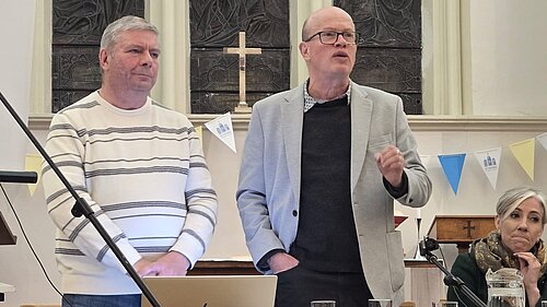 Councillors Tony Lillico and Paul de Kort are standing, whilst Paul addresses the room. Daisy Cooper MP is sat next to Paul, listening to his speech.