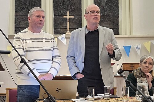 Councillors Tony Lillico and Paul de Kort are standing, whilst Paul addresses the room. Daisy Cooper MP is sat next to Paul, listening to his speech.