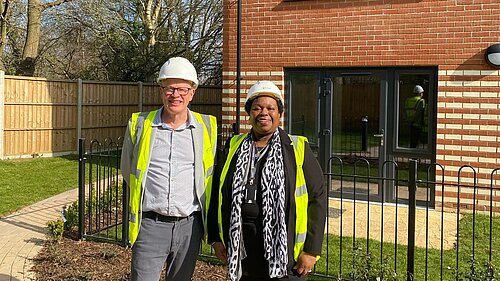 Councillors Paul de Kort and Jenni Murray are stood, both wearing hard hats and hi-vis vests, in front of one of the homes in The Hedges.