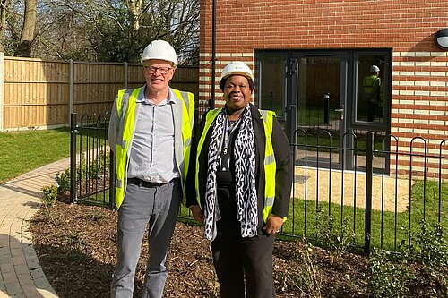 Councillors Paul de Kort and Jenni Murray are stood, both wearing hard hats and hi-vis vests, in front of one of the homes in The Hedges.