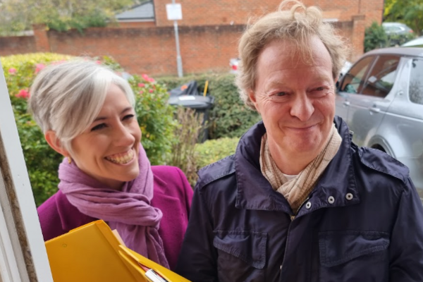 Chris and Daisy are standing on a doorstep, holding clipboards and smiling.